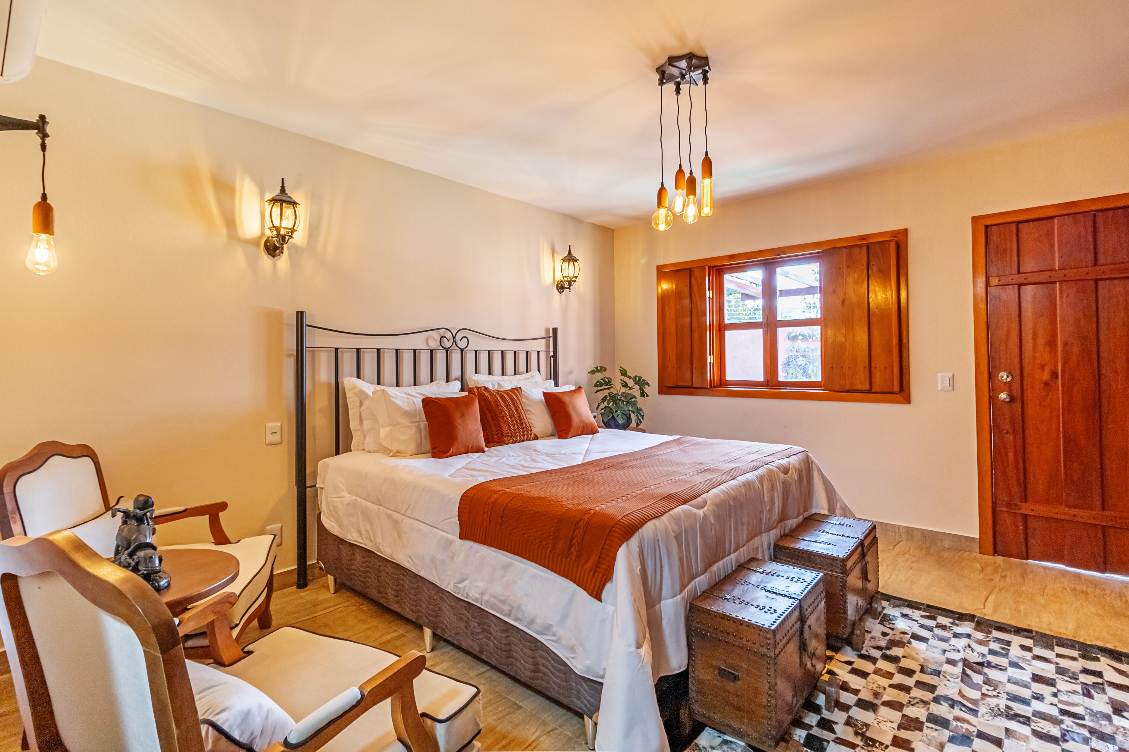 Un dormitorio con una cama grande, cabecero de madera, mesitas de noche, una silla, una pequeña mesa, una ventana con contraventanas de madera y un suelo de cuadros.