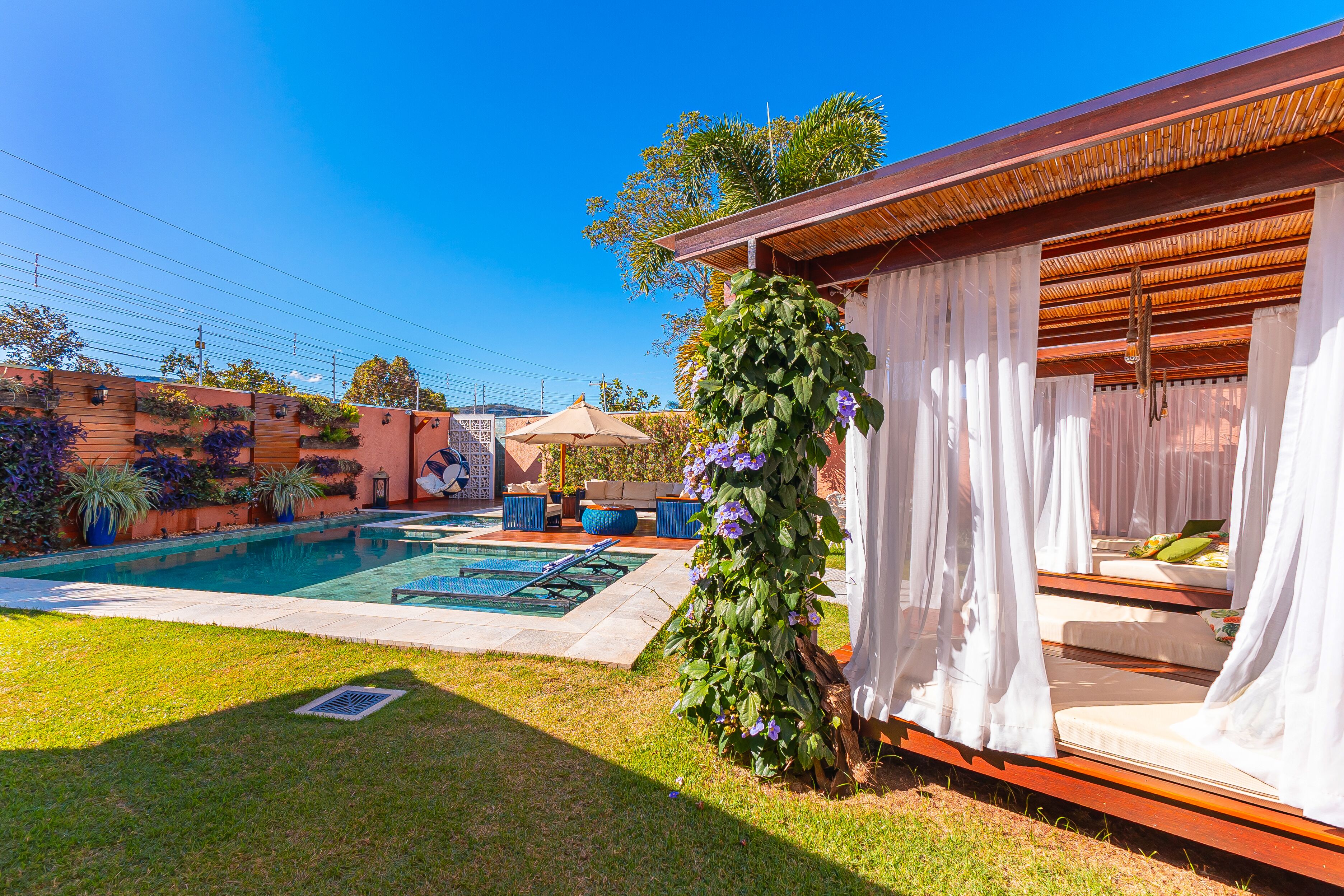 Un área de piscina con muelle de madera, zona de estar cubierta y pérgola con plantas trepadoras.