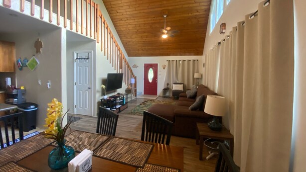 A spacious living area with a wooden ceiling, a television, a sofa, and a dining table with placemats. - Charming AFrame home w/2BR, 2BA, office suite, AC, WiFi & outdoor living (Keaau)