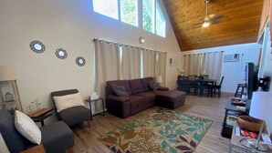 A spacious living room with a brown sectional sofa, a grey armchair, and a patterned rug. A dining area with a table and chairs is visible in the background. - Charming AFrame home w/2BR, 2BA, office suite, AC, WiFi & outdoor living (Keaau)