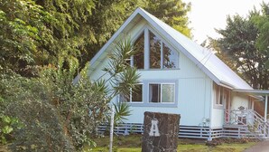 A light blue house with a stone well, surrounded by lush greenery. - Charming AFrame home w/2BR, 2BA, office suite, AC, WiFi & outdoor living (Keaau)