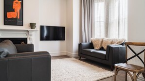 A living room with a fireplace, a flat-screen TV mounted on the wall, a grey sofa, a wooden chair, and a large window with curtains. - Premium Living on Stuart Street (Dunedin)