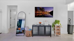 Un salon moderne avec un tableau représentant un paysage urbain, un grand miroir, un meuble gris, des paniers en osier et une plante en pot.