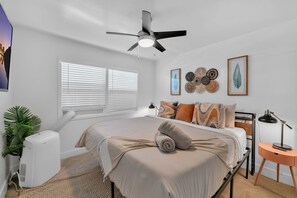 A bedroom with a bed, a ceiling fan, a heater, a bedside table with a lamp, and decorative wall art.