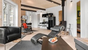 A modern living room with a sofa, two chairs, a dining table with a fruit bowl, and a black bookshelf.