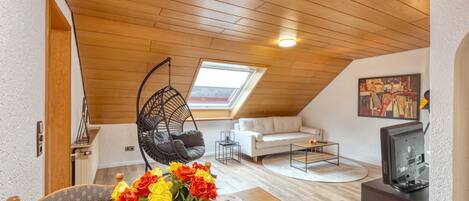A cozy loft apartment with a wooden ceiling, a hanging chair, a white sofa, a dining table with a vase of flowers, and a television.