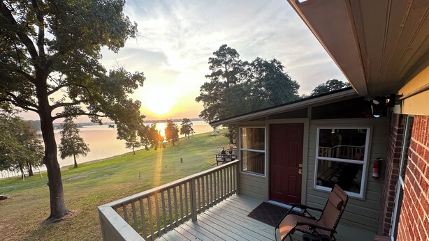 A wooden deck with two chairs and a table, overlooking a lake with trees and a house in the background. - Lake O' The Pines Lake Front
Two Bedroom Home - A Slice of Paradise (Jefferson)