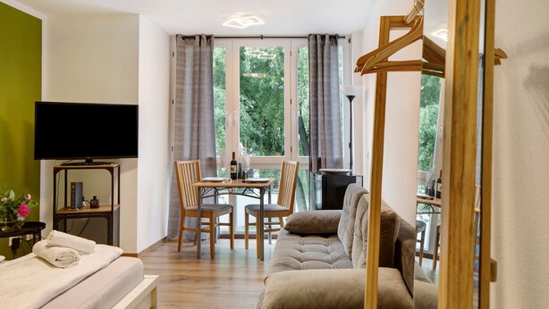 Ein Zimmer mit einem Bett, einer Couch, einem Esstisch mit Stühlen, einem Fernseher und einem Fenster mit Vorhängen.