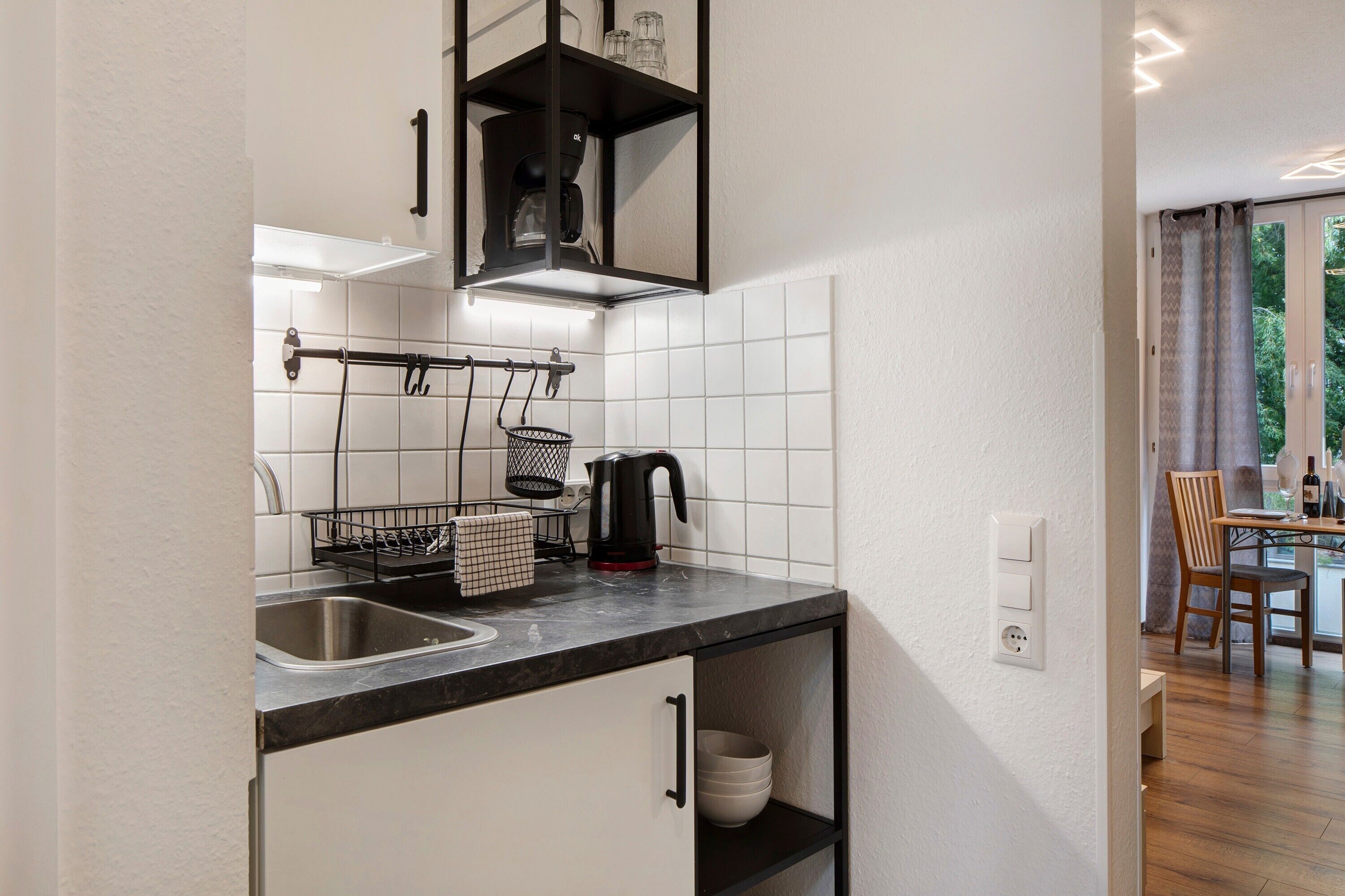 A modern kitchen with white tiles, a black countertop, a coffee machine, and a dining area with a table and chairs.