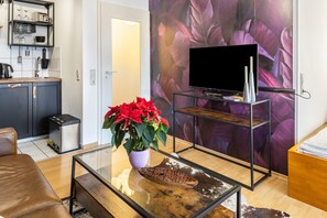 Ein Wohnzimmer mit Fernseher auf einem Schrank, einem Couchtisch mit Pflanze, einer Couch und einer Küchenzeile im Hintergrund.