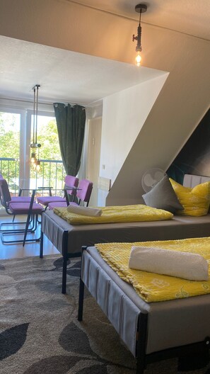 Ein Zimmer mit zwei Betten, einem Tisch mit Stühlen, einer Pendelleuchte und Blick auf Grünfläche vor dem Fenster.