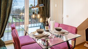 A dining area with a glass table, red upholstered chairs, a hanging light fixture, and a view through a large window.