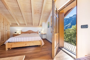 Un dormitorio con techo de madera, una cama con colcha a rayas, lámparas de noche, una mesita de noche y vistas a un balcón con montañas.