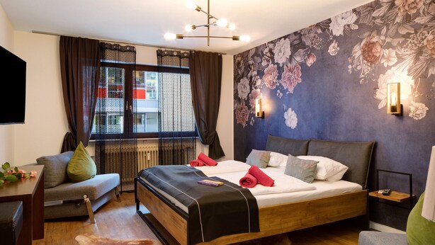 A bedroom with a bed, a sofa, a desk, a radiator, a window with curtains, and a floral wall. - Holiday Apartment 'Am Bahnhof Freiburg' with Wi-Fi (Freiburg im Breisgau)