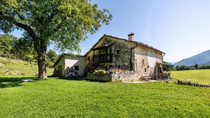 A rustic stone house with a balcony, surrounded by a lush green field and a large tree.