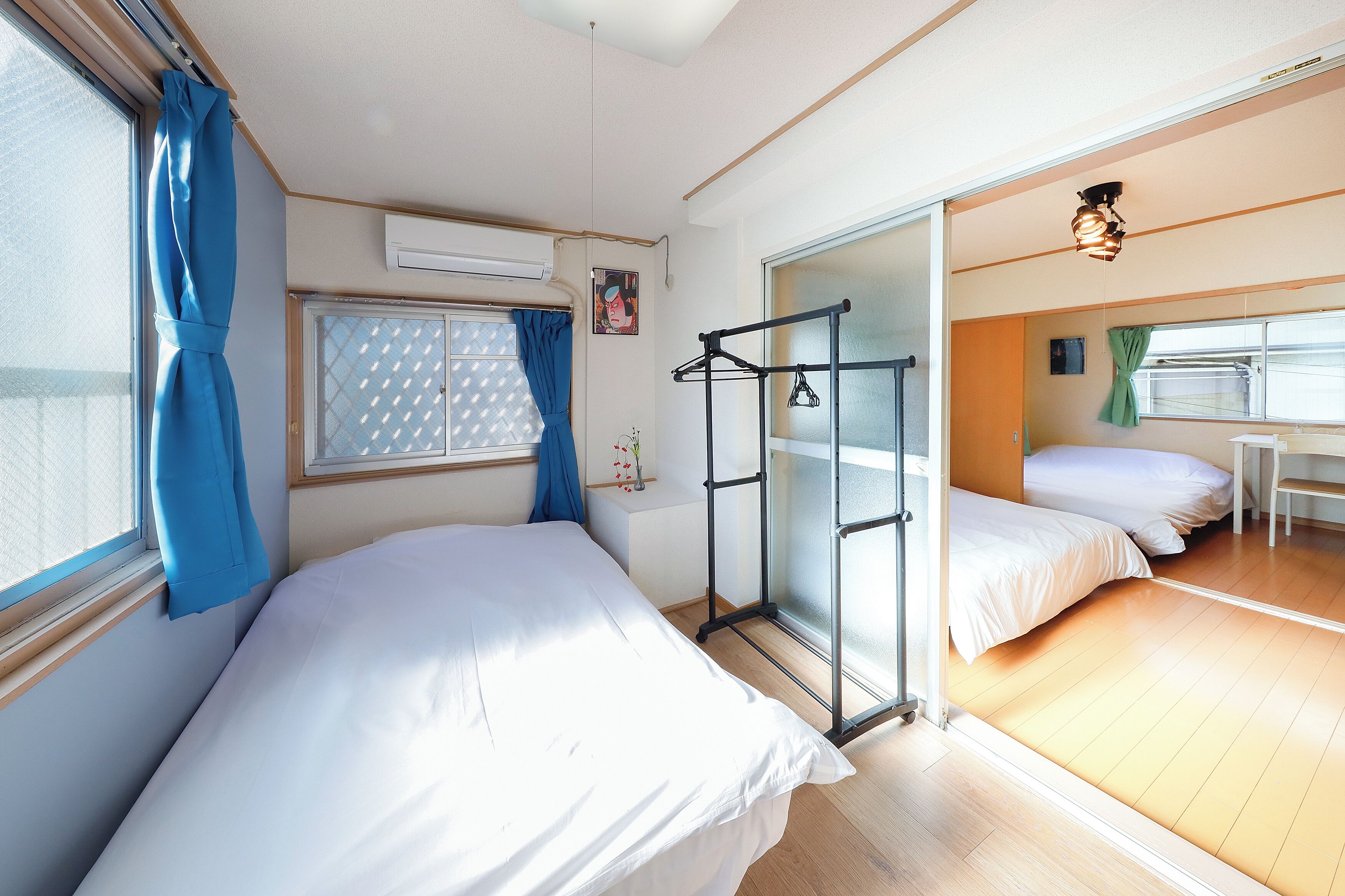 A room with two beds, a window with blue curtains, a drying rack, and a small table with a vase.