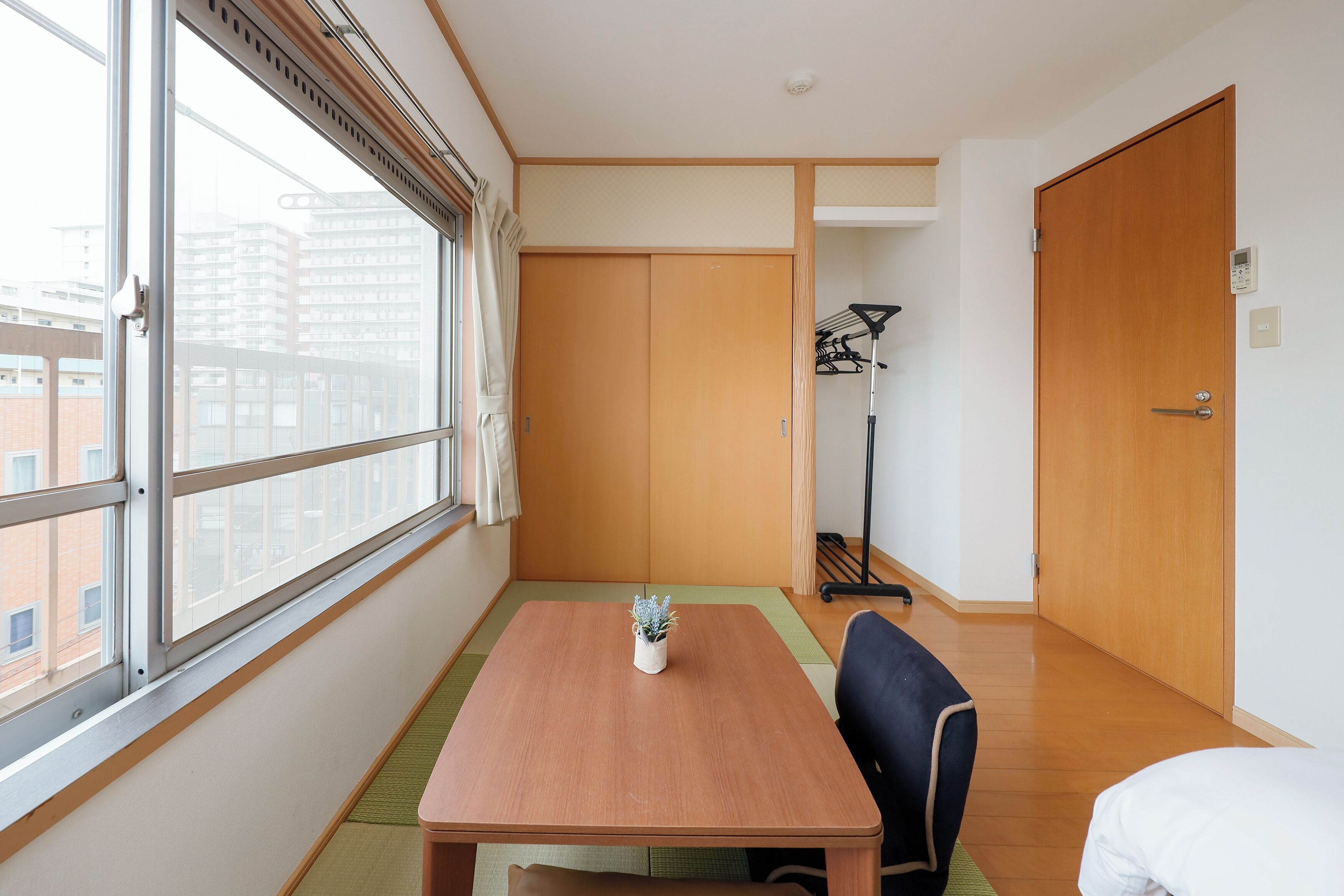 A hotel room with a large window, a wooden table, a chair, a bed, and a wardrobe.