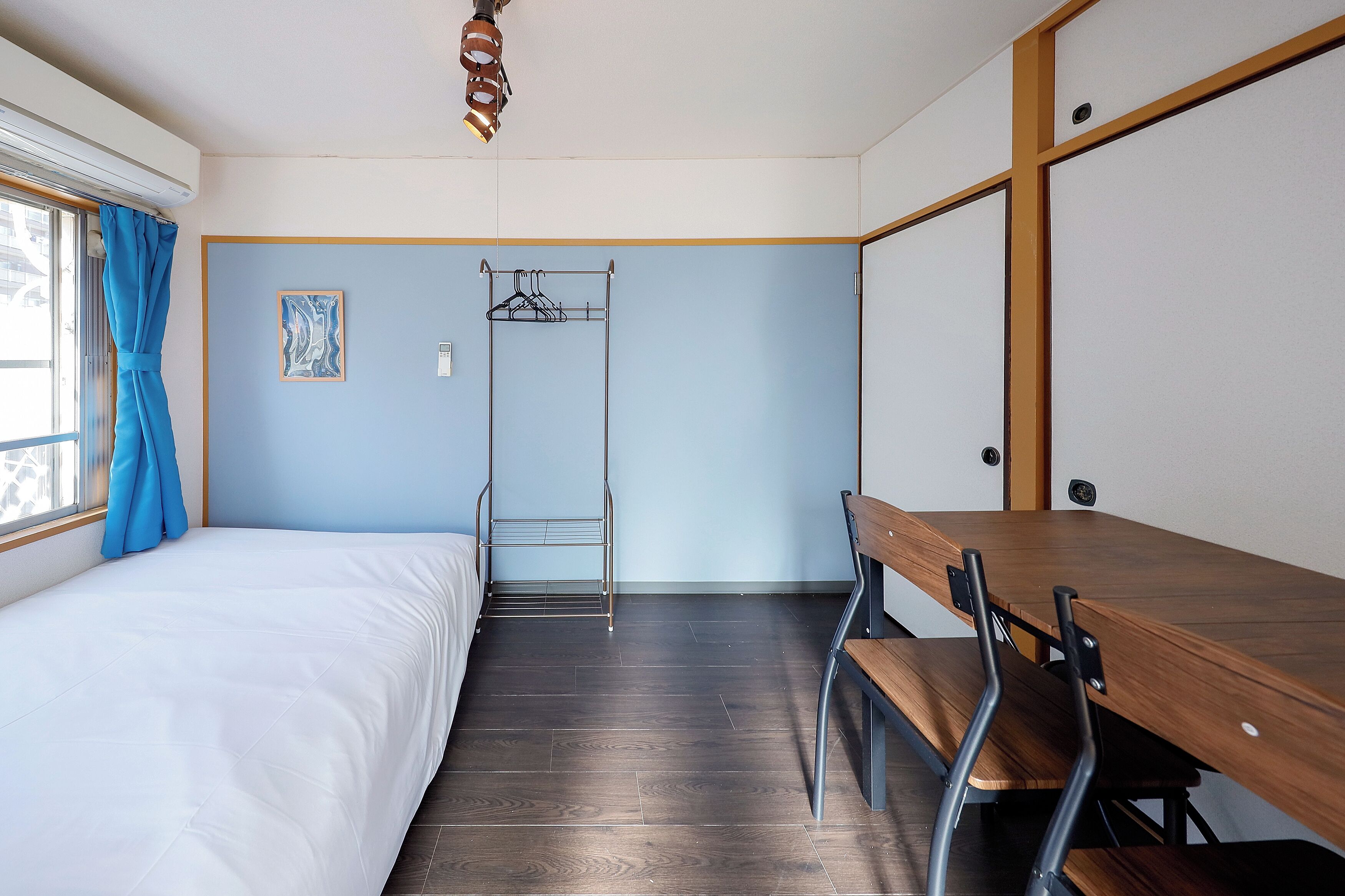 Een kamer met een bed, een houten tafel met stoelen, een kledingrek en een raam met gordijnen.