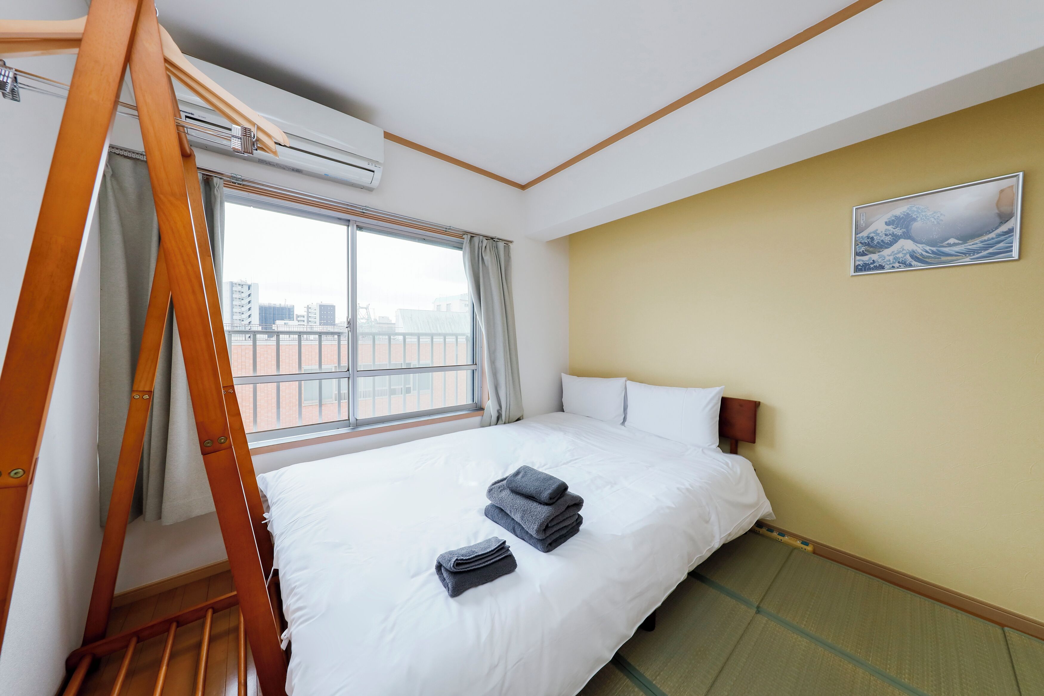 A hotel room with a bed, a wooden ladder, an air conditioning unit, a window with curtains, a tatami mat, and a painting on the wall.