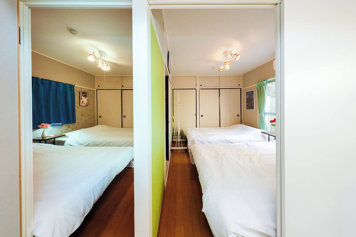 A hotel room with two beds, a wooden floor, a bathroom with a shower curtain, and a window with curtains.