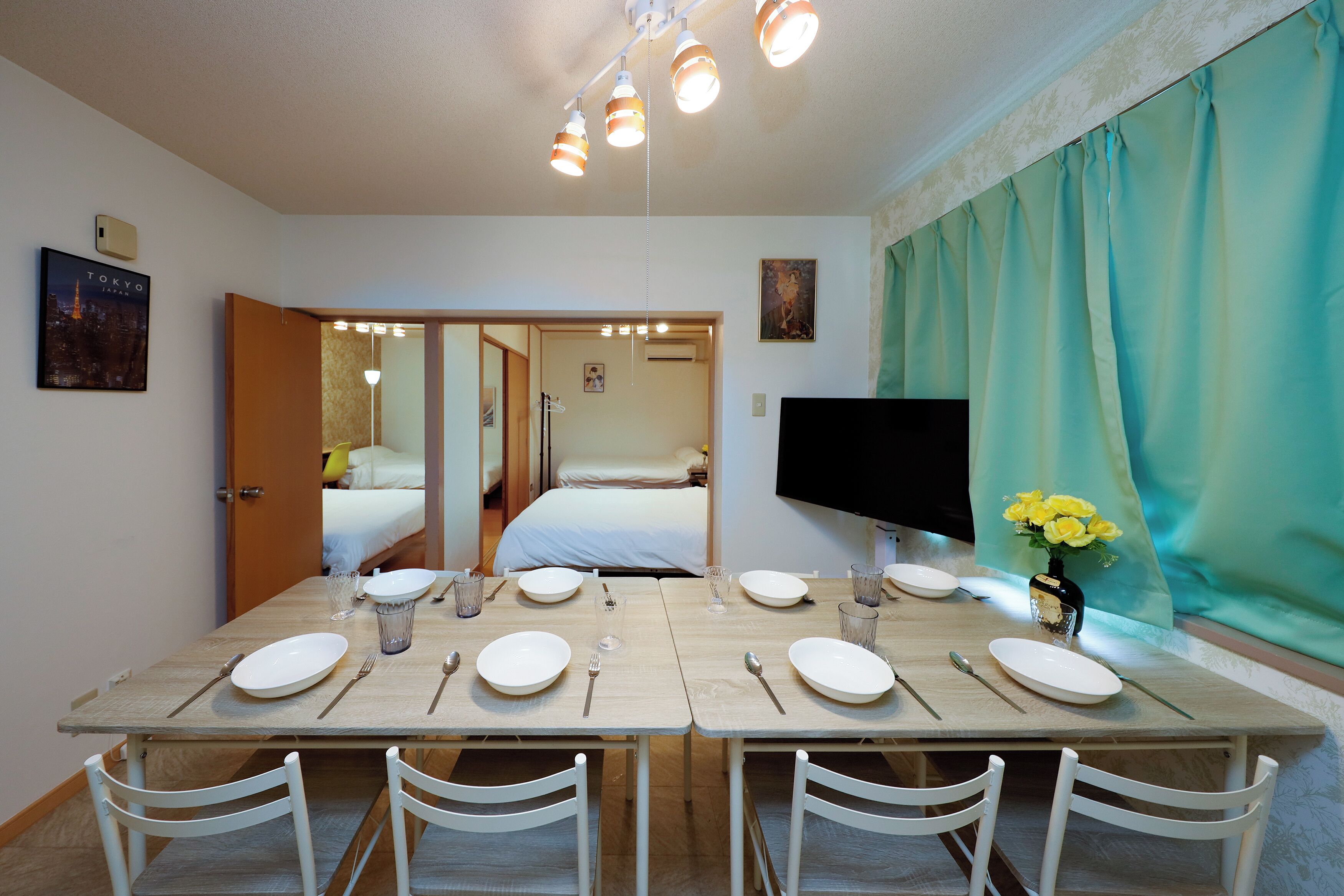A dining area with a table set for four, a bed in the background, a TV on the wall, and a window with curtains.