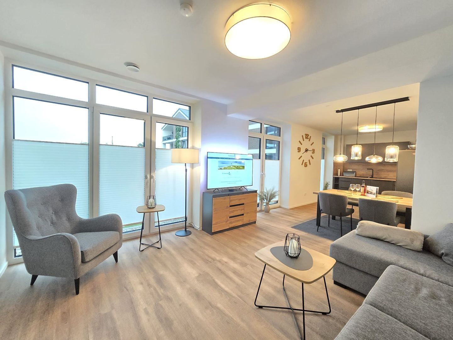 Een moderne woonkamer met een bank, een salontafel en een televisie. Er is een eethoek met een tafel en stoelen. De kamer heeft grote ramen en een keuken met een eettafel en stoelen.