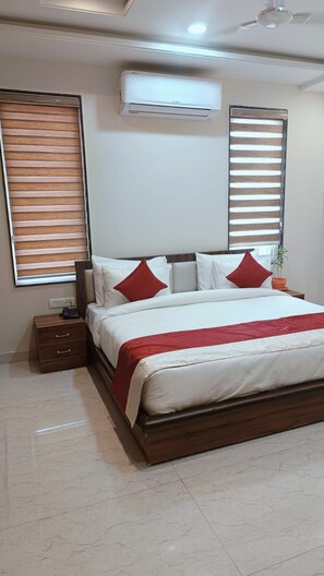 A modern bedroom with a large bed, a bedside table, an air conditioning unit, and wooden blinds. - Hotel Wings Grand (Alwar)