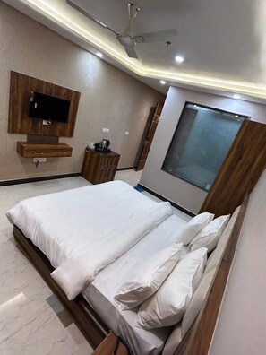 A modern bedroom with a large bed, a ceiling fan, a mounted TV, and a window. - Hotel Wings Grand (Alwar)
