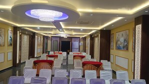 A hotel conference room with rows of white chairs, red tables, and a large chandelier. - Hotel Wings Grand (Alwar)
