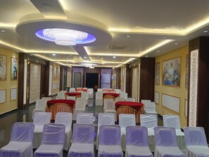 A hotel conference room with rows of white chairs, red tables, and a large chandelier. - Hotel Wings Grand (Alwar)