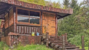 Ein rustikales Holzhaus mit großem Fenster und Tür, umgeben von viel Grün und einem Holzzaun.