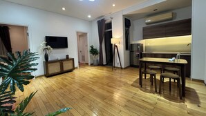 A spacious living area with a wooden floor, a dining table with chairs, a flat-screen TV mounted on the wall, and a modern kitchen with an air conditioning unit.
