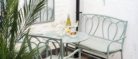 A metal table and chairs set against a white wall, with a bottle and glasses on the table.