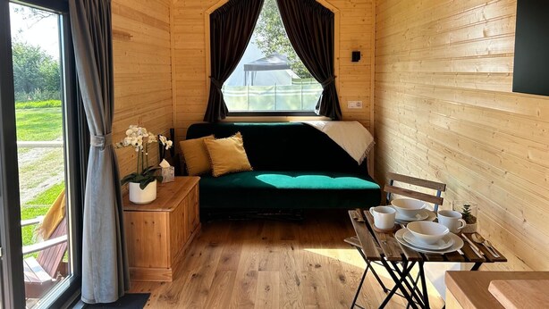 Ein gemütliches Blockhaus-Interieur mit einem grünen Sofa, Holzwänden, einem Esstisch mit Tellern und Besteck sowie einem Fenster mit Vorhängen.
