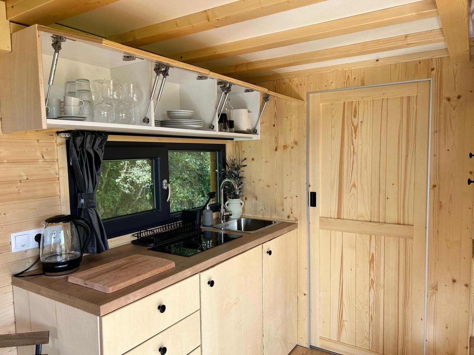 Een kleine keuken met houten keukenkastjes, een ingebouwde kookplaat en een raam met gordijn.