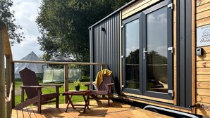 Eine kompakte, moderne Hütte mit einer Holzdielen-Terrasse, einer überdachten Sitzgelegenheit und einem großen Schiebeglasfenster.