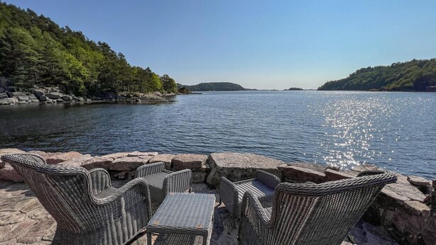Vista al agua con muebles de mimbre y una mesa de piedra, con vista a un lago tranquilo y colinas boscosas.
