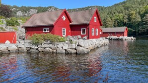 Una casa roja de madera con techo de tejas, ubicada en una orilla rocosa junto a un lago tranquilo, rodeada de vegetación y colinas.