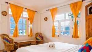 A bedroom with a large bed, two wicker chairs, a bench, and windows with yellow curtains. - Oak Land House (Manali)