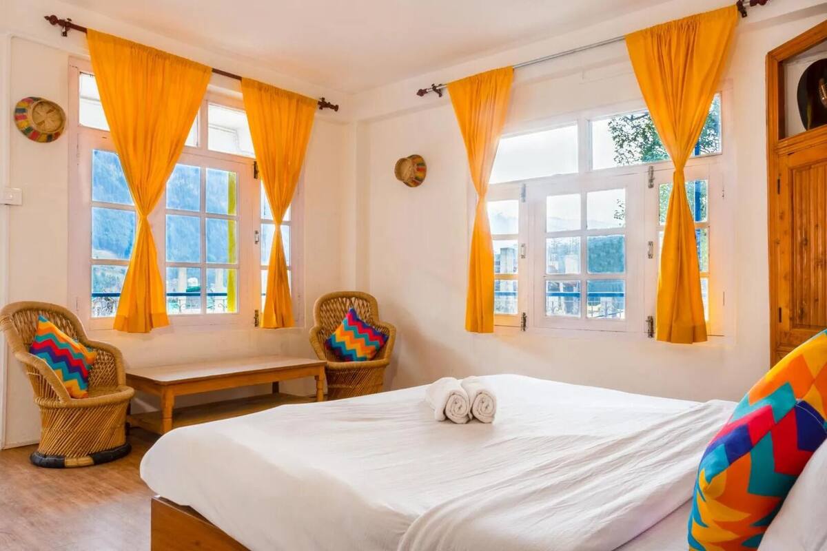 Une chambre à coucher avec un grand lit, deux fauteuils en osier, un banc et des fenêtres ornées de rideaux jaunes.