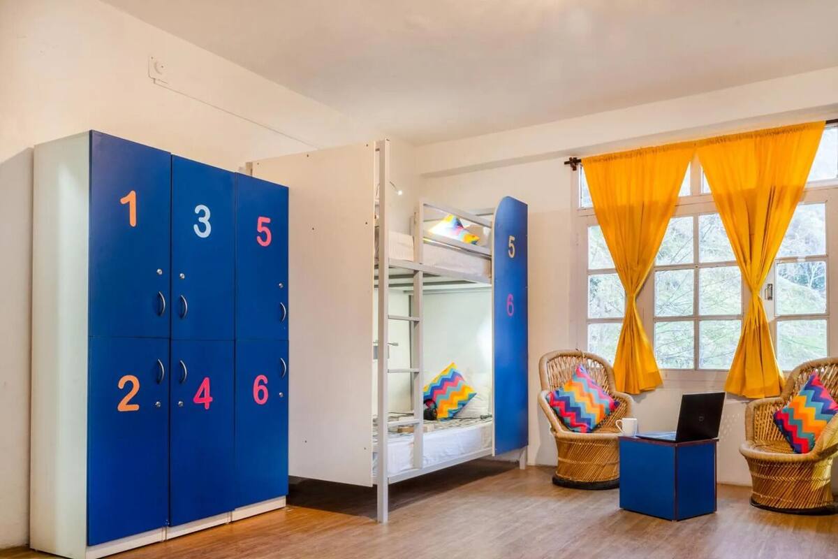 Une chambre avec des lits superposés, une armoire avec des compartiments numérotés, des chaises en osier et une fenêtre avec des rideaux jaunes.