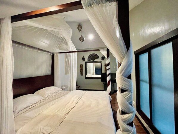 A four-poster bed with white linens and a canopy, a large window with blinds, and a mirror on the wall.