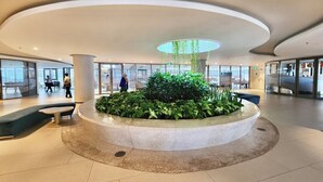 A spacious lobby with a central circular seating area surrounded by greenery, large windows, and a modern ceiling design.