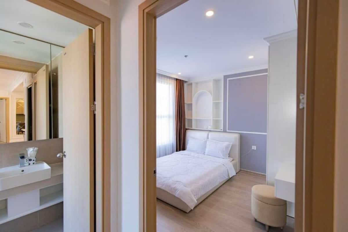 Quarto moderno com cama grande, mesa de cabeceira, espelho e um pequeno banquinho.