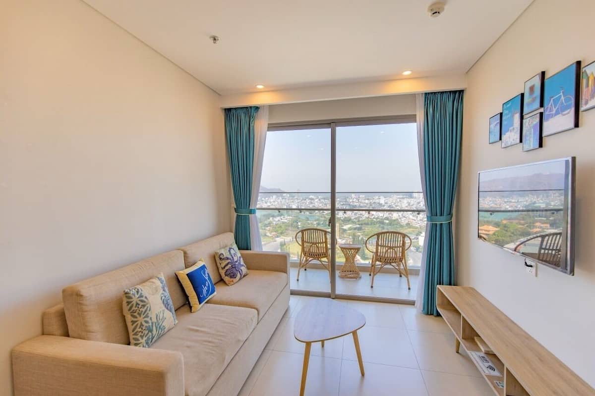 Uma sala de estar moderna com um sofá, uma mesa de centro e duas cadeiras de vime. Uma grande janela oferece vista para a cidade.