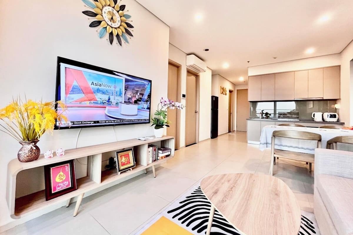 Uma sala de estar moderna com televisão de tela plana, um relógio de parede decorativo, um espaço de refeições com mesa e cadeiras, e uma cozinha com armários e eletrodomésticos.