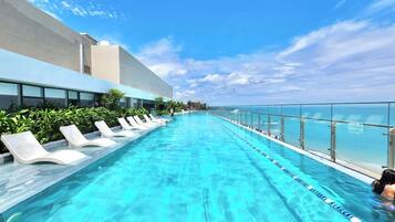 A long, clear swimming pool with white lounge chairs on one side and a glass railing on the other.