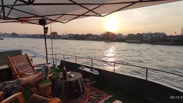 A boat deck with a table, chairs, and a view of the sunset over a river.