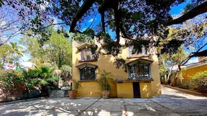 Um edifício amarelo com um pátio pavimentado, rodeado por árvores e plantas em vasos.