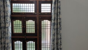 A room with a large window featuring a wooden frame and metal bars, flanked by patterned curtains. - Rameshwaram Stay (Faizabad)